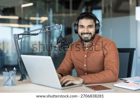 Young man smiling while broadcasting an online radio show or recording a podcast, speaking into a professional microphone and typing on a laptop in a modern office setup