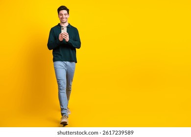 Young man smiling while checking his smartphone wearing casual green polo shirt on vibrant yellow background