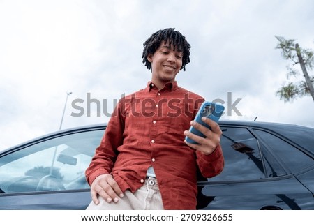 Young man smiling while checking his mobile phone leaning against a car
