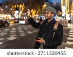 Young man smiling while hailing a taxi on a vibrant city street at night, holding a smartphone and wearing a backpack and hat, surrounded by blurred evening lights