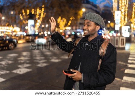 Young man smiling while hailing a taxi on a vibrant city street at night, holding a smartphone and wearing a backpack and hat, surrounded by blurred evening lights