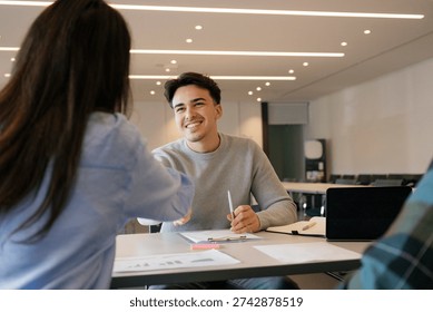 Young man smiling while shaking hands across a desk with a colleague in an office, celebrating a successful job interview, hiring decision or new professional partnership