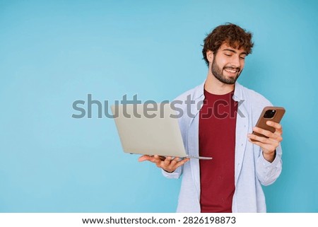 Young man smiling while simultaneously holding a laptop and smartphone, engaging with technology for business or leisure