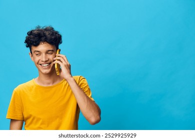 Young man smiling while talking on a smartphone against a bright blue background, wearing a yellow shirt, conveying joy and communication in a cheerful setting - Powered by Shutterstock