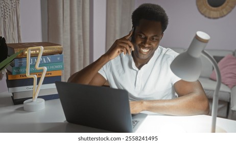 Young man smiling while talking on smartphone and working on laptop at home in cozy living room with books and lamp
