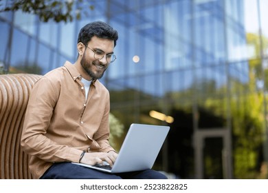 Young man smiling while using laptop outdoors in modern office environment demonstrating work flexibility and remote productivity. Business attire suggests professionalism and confidence - Powered by Shutterstock