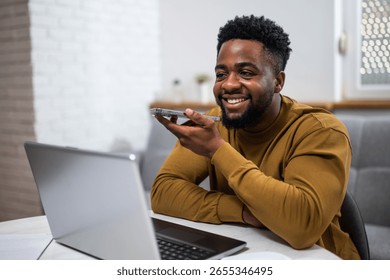 Young man smiling while using laptop and talking on smart phone in his living room, working remotely.