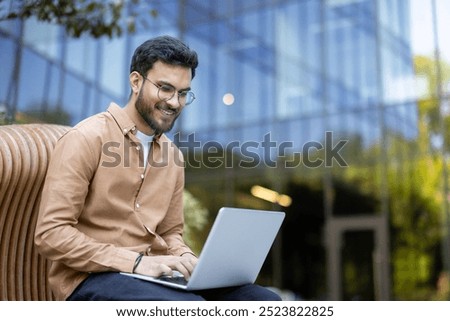 Young man smiling while using laptop outdoors in modern office environment demonstrating work flexibility and remote productivity. Business attire suggests professionalism and confidence