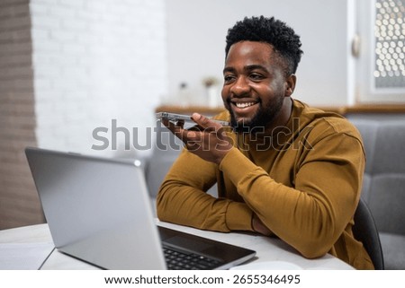 Young man smiling while using laptop and talking on smart phone in his living room, working remotely.