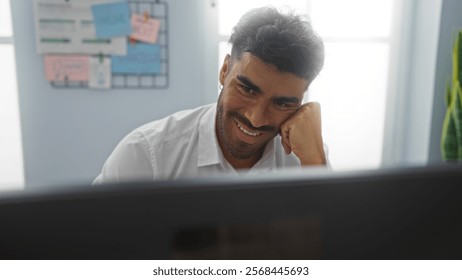 Young man smiling while working at a computer in an office interior, demonstrating a positive and productive work atmosphere.