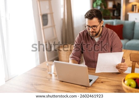 Young man smiling while working from home on his laptop. Cozy living room with bookshelves, warm lighting, indoor plants, and a bowl of fresh fruits, creating a balanced work environment