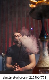 a young man smokes a hookah sitting on a sofa, a hookah pipe in his mouth, exhales white smoke. in the foreground a bowl of hookah on grapefruit