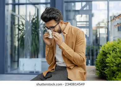 Young man sneezing and having a runny nose allergy sitting on a bench in the daytime outside an office building, hispanic businessman sick with a tissue near his nose. - Powered by Shutterstock