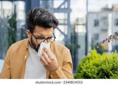 Young man sneezing and having a runny nose allergy sitting on a bench in the daytime outside an office building, hispanic businessman sick with a tissue near his nose.