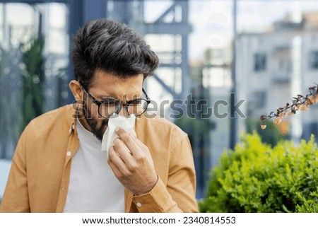 Young man sneezing and having a runny nose allergy sitting on a bench in the daytime outside an office building, hispanic businessman sick with a tissue near his nose.