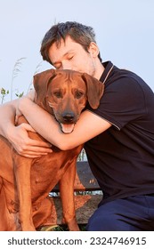 Young man snuggling and hugging his dog sitting outdoors. Close friendship, love and bond between owner and pet 