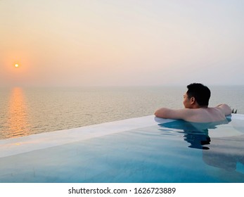 Young Man Soaking And Swimming In An Infinity Pool With Beautiful And Peaceful Ocean And Sunset View During His Holiday. Solo Travel For Weekend Getaway And Lifestyle Concept