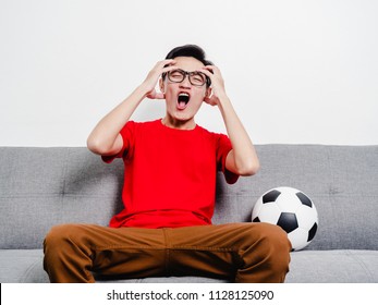 Young Man Soccer Fan Crying Face After Watching Football Match On Tv While Sitting On Grey Sofa And Wearing Red Shirt Short Sleeve.