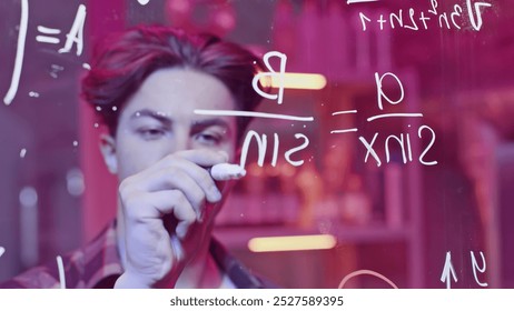 Young man solves mathematical equations on blackboard. Media. Young man solves complex equations on transparent board. Genius writes equations on blackboard in neon light