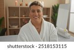 Young man in a spa room wearing a robe, smiling and enjoying the relaxing wellness ambiance with candles and plants in the background.