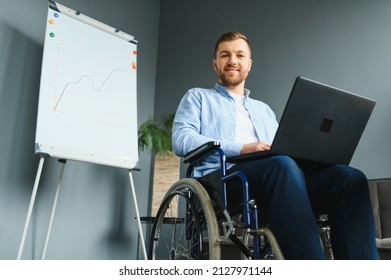 Young Man With Special Needs In Casual Clothes Working On Wireless Laptop. Male Freelancer Working From Home While Sitting In Wheelchair.