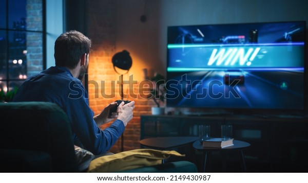 Young
Man Spending Time at Home, Sitting on a Couch in Stylish Loft
Apartment and Playing Arcade Car Video Games on Console. Male Using
Controller to Play Street Racing Drift
Simulator.