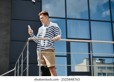 A young man, sporting a stylish striped sweater and shorts, stands on a balcony with his smartphone. - Powered by Shutterstock