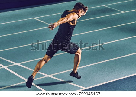 Young man sprinting on a blue indoor racetrack wearing sports clothing & looking tired after a training session.