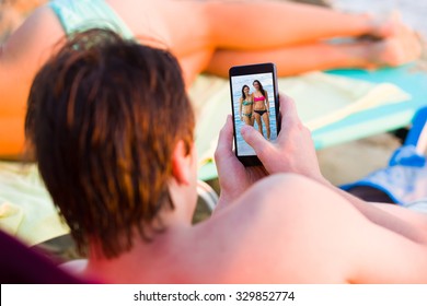 Young Man Stalking Girls On Facebook On The Beach Next To His Girlfriend.