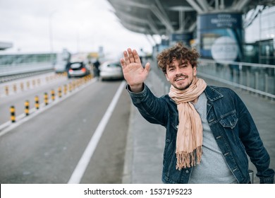 Young Man Stand Outside Of Airport Alone. Wave With Hand. Try To Catch Taxi Cab Or Transportation. Come Back From Trip Or Travel. Stand Alone And Wait For Car. Daylight