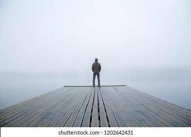 Young man standing alone on edge of footbridge and staring at lake. Mist over water. Foggy air. Early chilly morning in autumn. Beautiful freedom moment and peaceful atmosphere in nature. Back view.