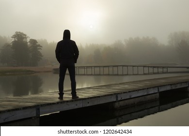 Young man standing alone on lake footbridge and staring at sunrise in gray, cloudy sky. Mist over water. Foggy air. Early chilly morning. Dark, scary moment and gloomy atmosphere. Back view. - Powered by Shutterstock