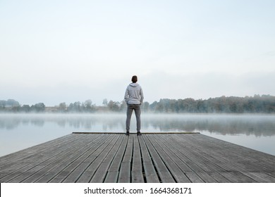 Young man standing alone on wooden footbridge and staring at lake. Thinking about life. Mist over water. Foggy air. Early chilly morning. Peaceful atmosphere in nature. Enjoying fresh air. Back view. - Shutterstock ID 1664368171