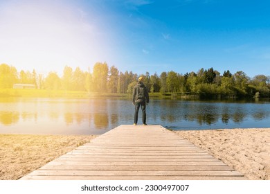 Young man standing alone on edge of wooden footbridge and staring at lake.  Peaceful atmosphere in nature. Enjoying fresh air in sunny spring day. Back view. - Shutterstock ID 2300497007