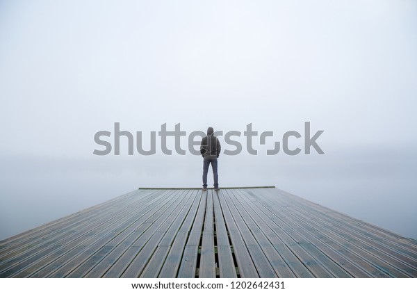 Young man standing alone on edge of footbridge and\
staring at lake. Mist over water. Foggy air. Early chilly morning\
in autumn. Beautiful freedom moment and peaceful atmosphere in\
nature. Back view.