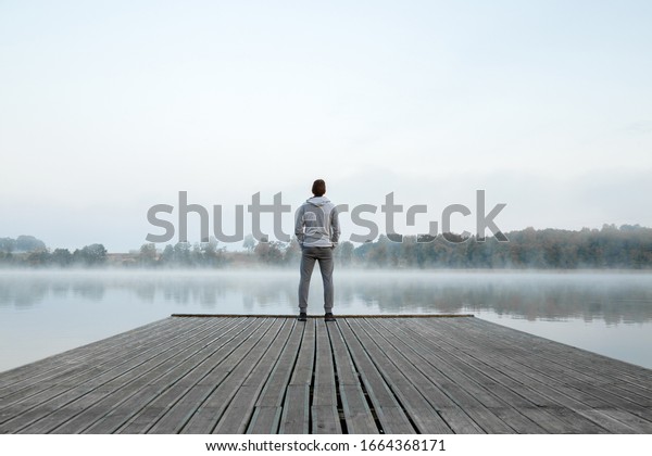 Young man standing alone on wooden footbridge and\
staring at lake. Thinking about life. Mist over water. Foggy air.\
Early chilly morning. Peaceful atmosphere in nature. Enjoying fresh\
air. Back view.