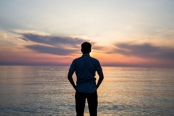 Young Man Standing At The Beach In Front Of Amazing Sea View At Sunset Or Sunrise And Thinking About His Future. Rear View