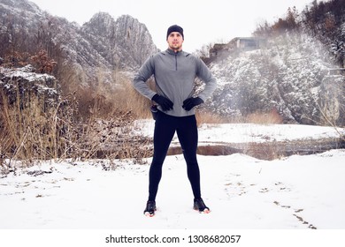 Young Man Standing By The River In Winter Day. Marathon Ultra Trail Runner Resting By The River