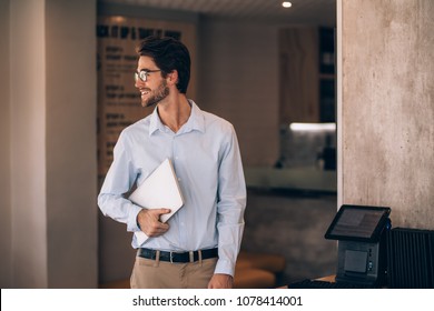 Young Man Standing In A Cafe And Looking Away. Male Restaurant Manager With A Laptop.