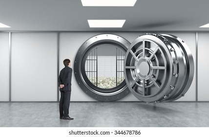 Young Man Standing In Front Of A Big Unlocked Round Metal Safe In A Bank With Lock-boxes And Heaps Of Dollars Inside, In A Depository, With His Back Half Turned Looking Inside, A Concept Of Aspiration