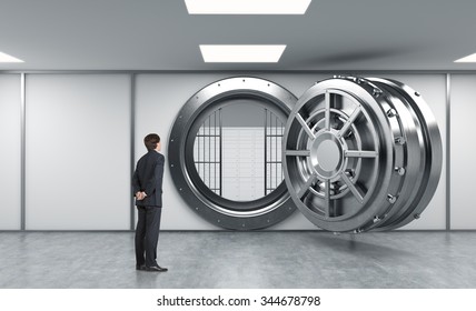 Young Man Standing In Front Of A Big Unlocked Round Metal Safe In A Bank With Lock-boxes Inside In A Depository With His Back Half Turned Looking Inside,  A Concept Of Aspirations