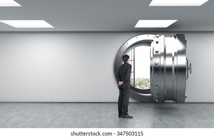 Young Man Standing In Front Of A Big Unlocked Round Metal Safe In A Bank With Heaps Of Dollars Inside, In A Depository, With His Back Half Turned Looking Inside,  A Concept Of Aspirations