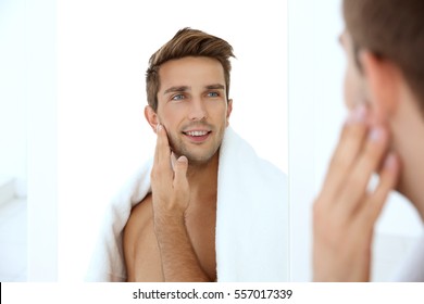 Young man standing in front of mirror and touching his face