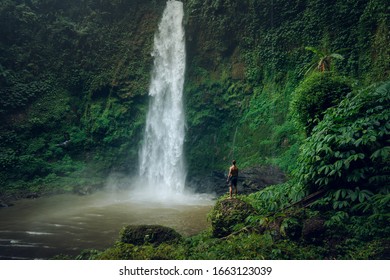 Young man standing in front of powerful Nungnung waterfall. Fun at the waterfalls in Bali. Concept about wanderlust traveling and wilderness culture
