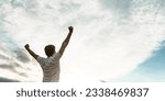Young man standing with his arms raised high in triumph under white cloudy sky with plenty of copy space.