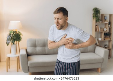 A young man is standing at home and holding his chest. Feels severe pain, shortness of breath, panic attack, heart attack, stroke. - Powered by Shutterstock