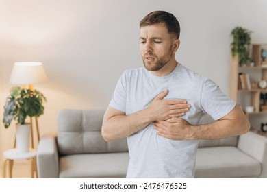 A young man is standing at home and holding his chest. Feels severe pain, shortness of breath, panic attack, heart attack, stroke. - Powered by Shutterstock