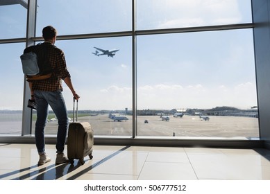 Young man is standing near window at the airport and watching plane before departure. He is standing and carrying luggage. Focus on his back