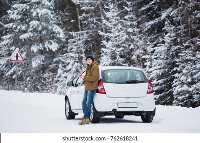 Young Man Is Standing Next To Car On A Winter Road. Cold Weather, Snow-covered Trees In The Winter Forest. Man Looking At The Camera And Holds Mobile Phone In His Hands. Winter Holidays. 