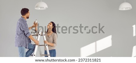 Young man standing on the ladder changing light bulb in the room with his smiling wife. Happy couple installing the chandelier in their new empty apartment. Electricity replacing concept. Banner.